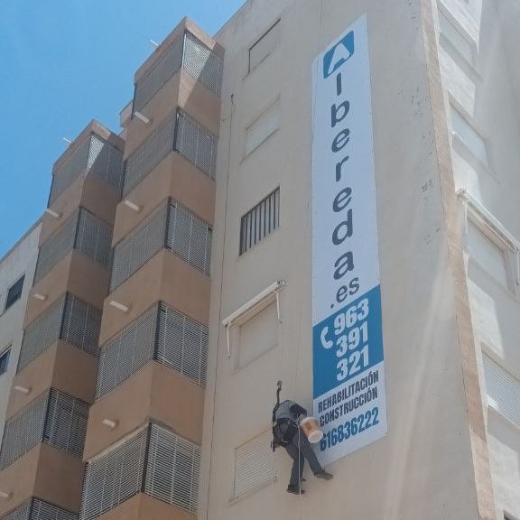 Trabajos verticales Obrero colgado con arneses en la fachada de un edificio junto con un cartel de Albereda con los teléfonos